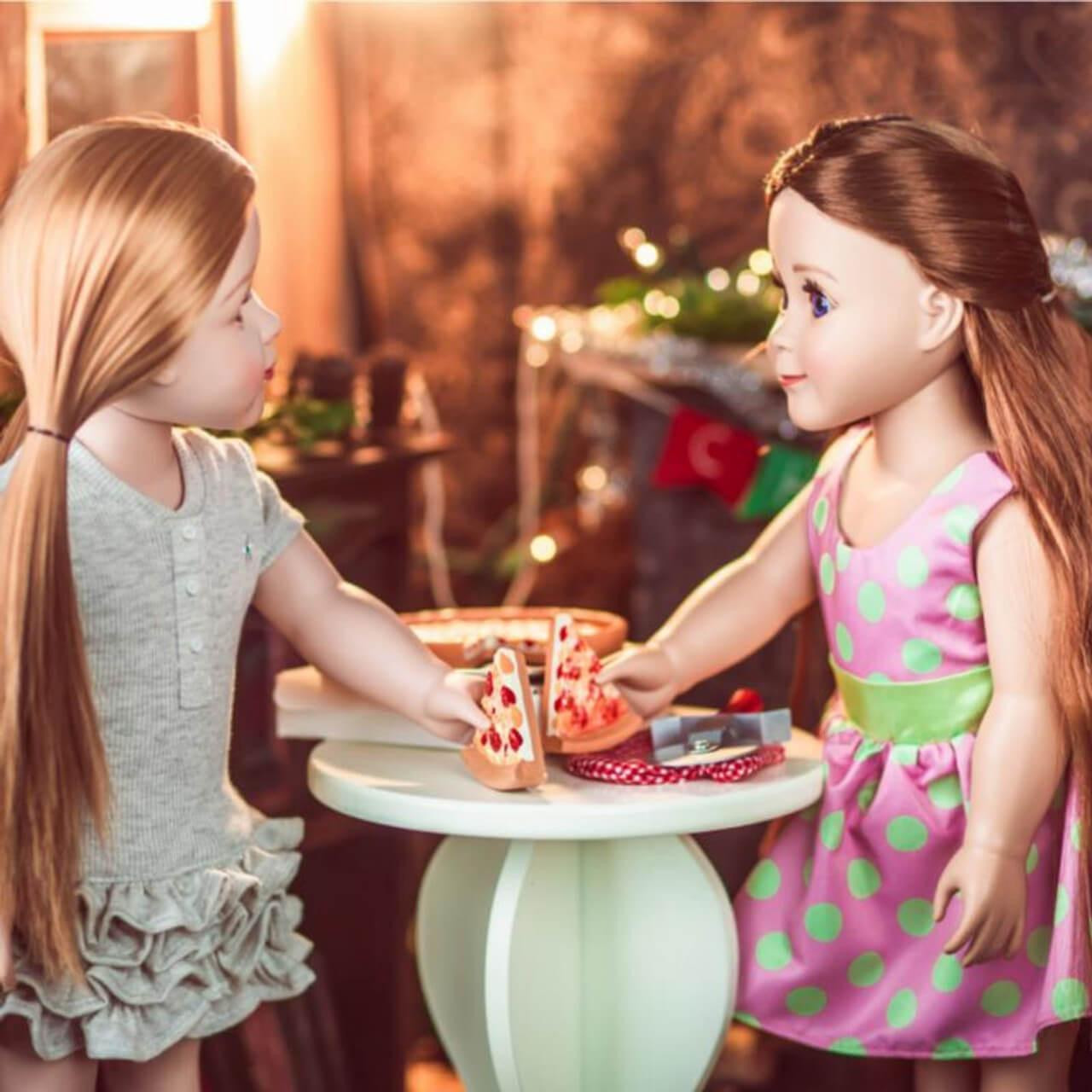 In home play scene two 18 inch girl dolls hold slices of pizza sitting at an 18" doll table.