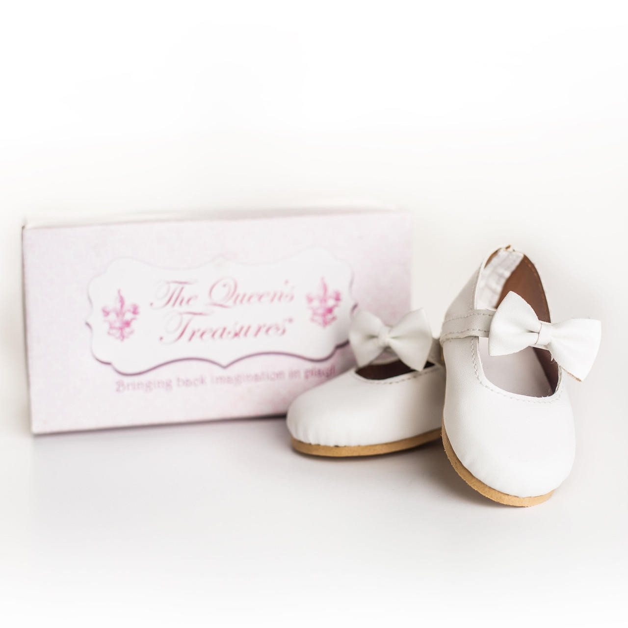 A pair of miniature white Mary Jane-style doll dress shoes stands next to their original packaging box against a solid white background. The shoe on the right faces forward, highlighting a prominent white bow on its ankle strap, while the second shoe sits slightly behind it. The rectangular box features a light pink lid printed with a decorative white label reading "The Queen's Treasures.".