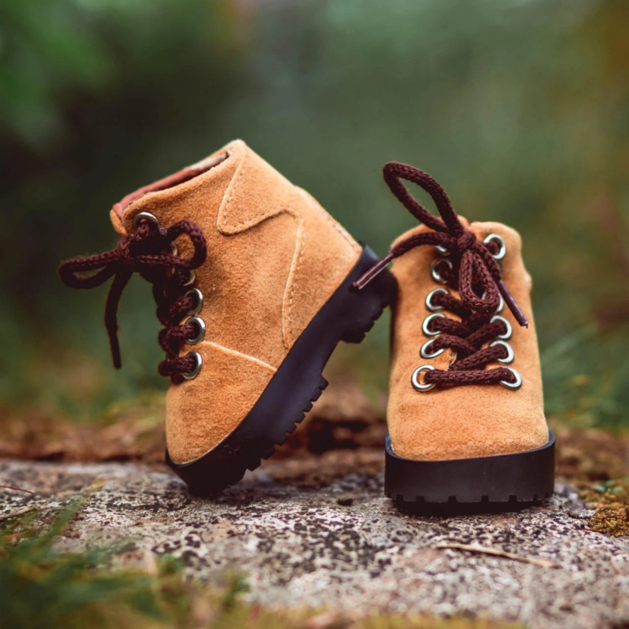 A pair of miniature tan suede doll hiking boots sits outdoors on a textured stone surface with a blurred green background. The boot on the left leans at a slight angle against the right boot, showcasing dark brown laces threaded through silver metal eyelets and thick, black rugged rubber soles with heavy treading.