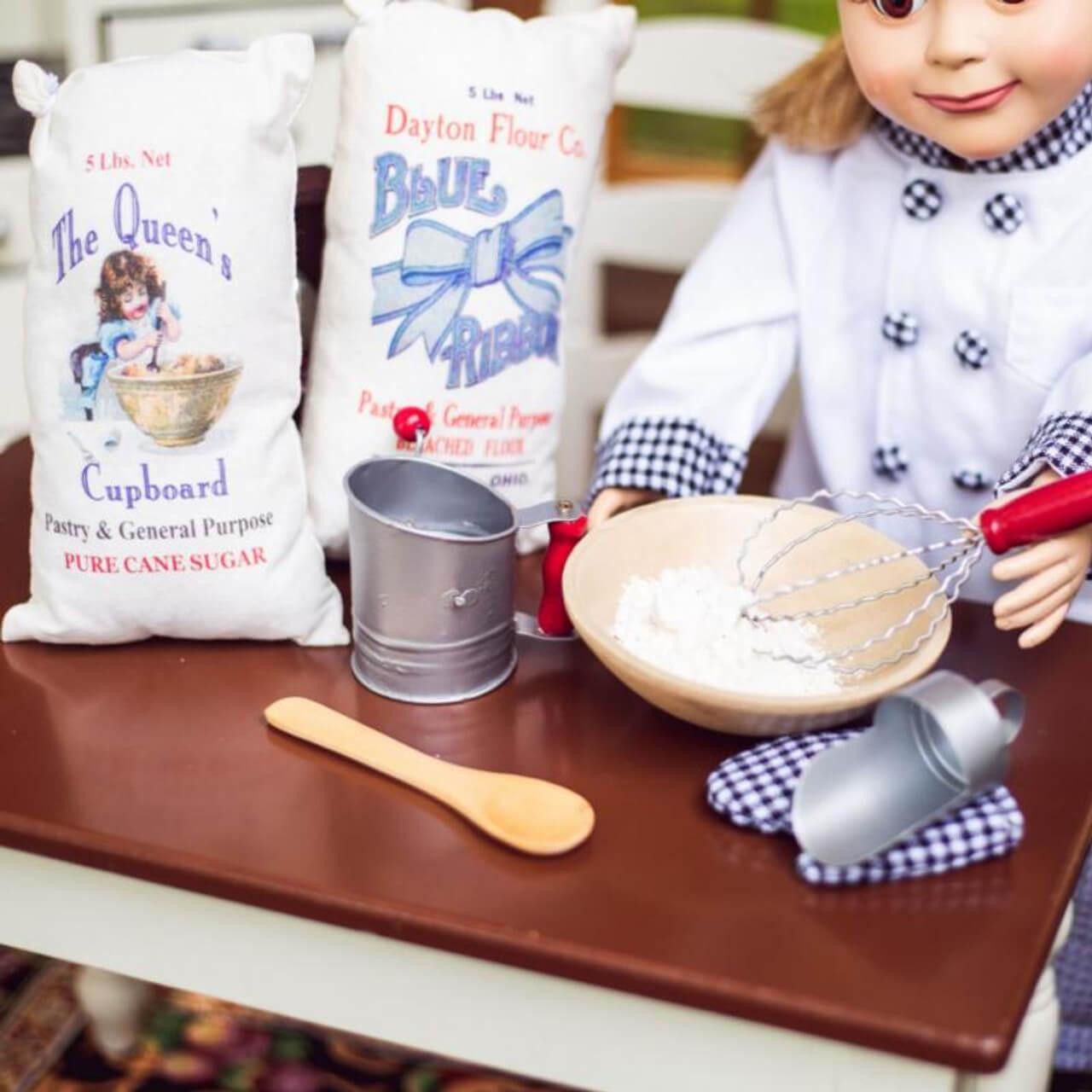 An 18-inch doll in a baker's jacket stirs a bowl of flour on a wooden table surrounded by miniature baking tools, a flour sifter, a scoop, and two fabric ingredient sacks by The Queen's Treasures®.