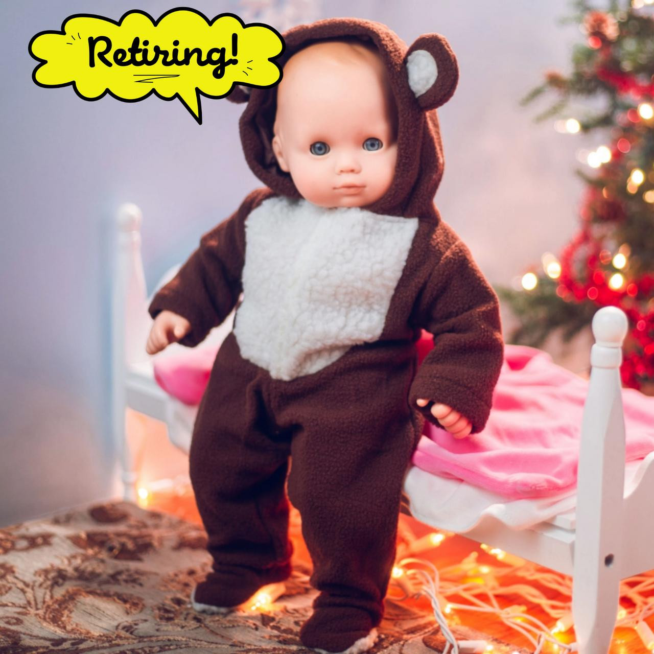 A baby doll wears a brown and white hooded bear onesie next to a white toy bed, with a lit Christmas tree in the background and a yellow "Retiring!" speech bubble.