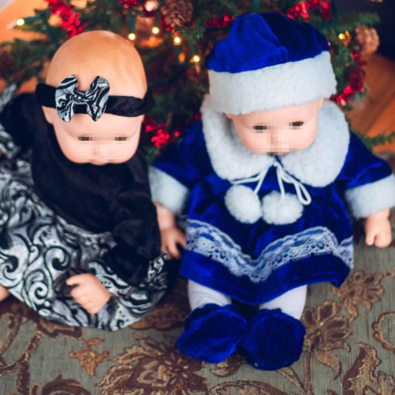 Two baby dolls sit together in front of a decorated Christmas tree; one wears a royal blue velvet holiday dress with matching hat, while the other wears a black and silver patterned dress with a bow headband.
