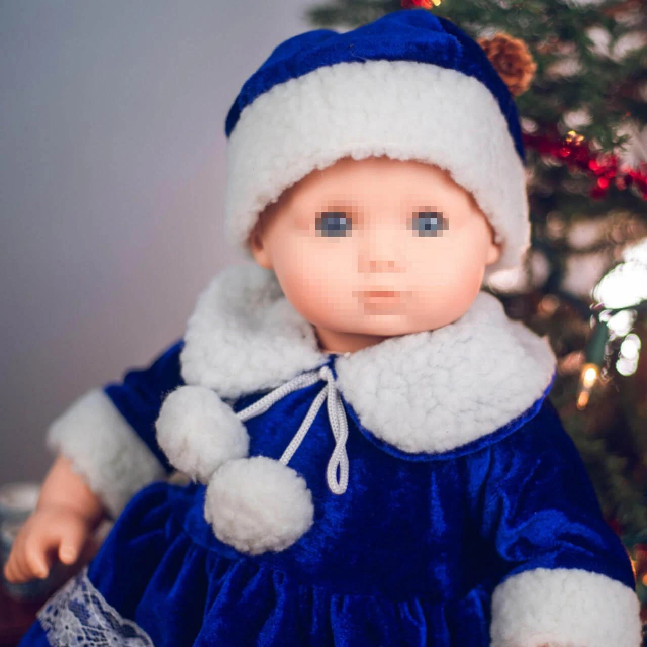 A close-up view of a baby doll with blue eyes wearing a royal blue velvet winter dress with plush white faux fur collar trim and a matching hat, set against a softly blurred Christmas tree background.
