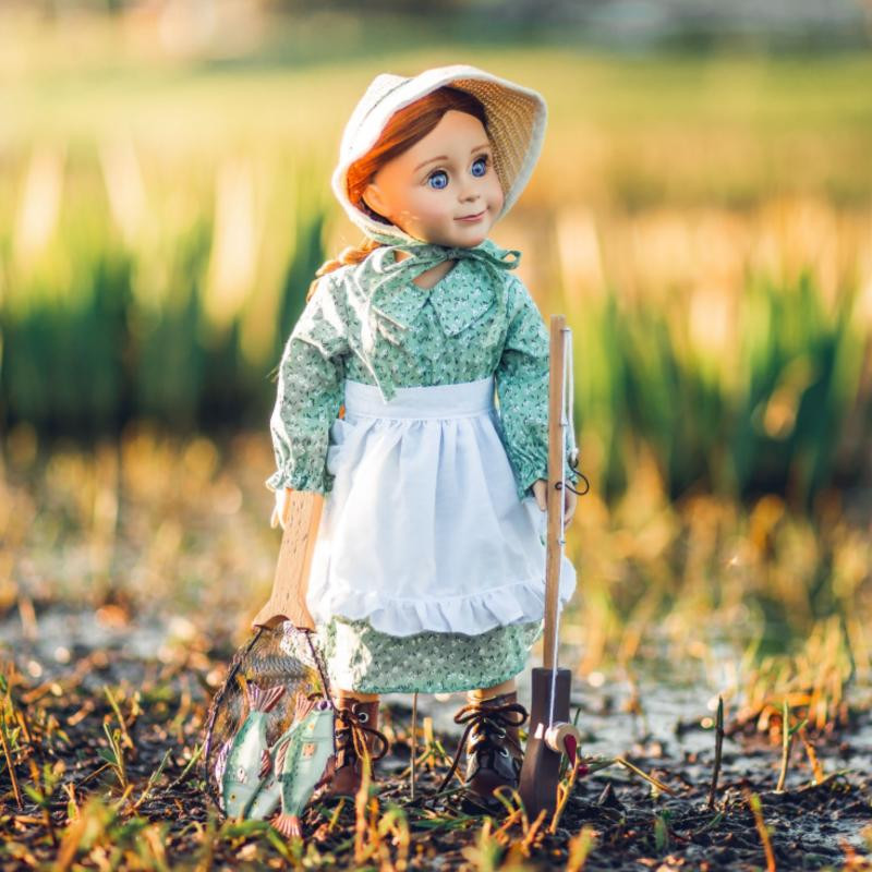 A  doll stands outdoors in a green floral dress, white apron, bonnet, and lace-up boots, holding a miniature wooden fishing pole and a net with fake fish.
