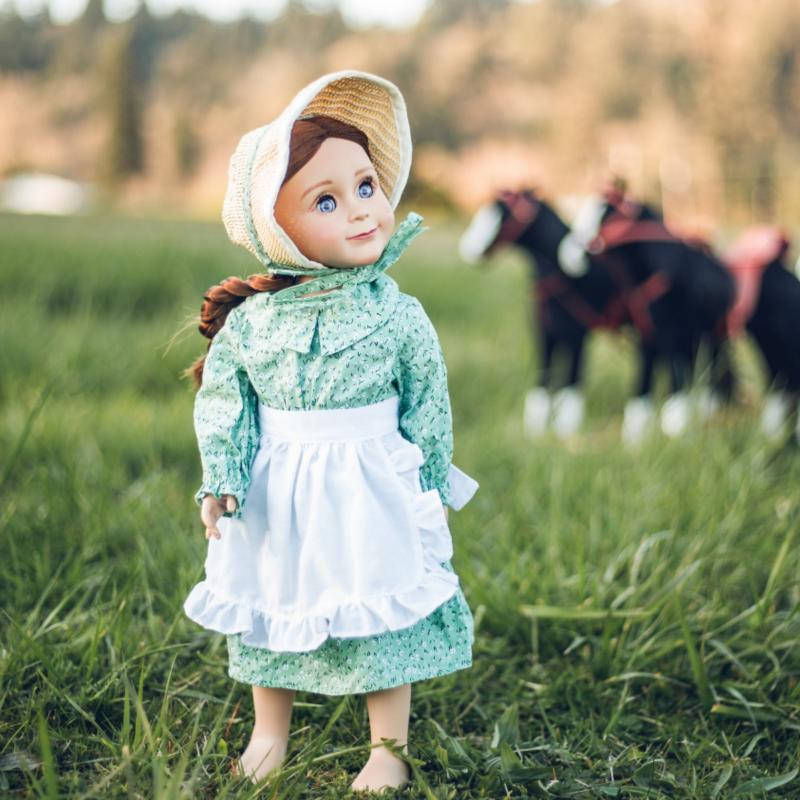 A pioneer doll in a green calico dress, white ruffled apron, and fabric bonnet stands in a grassy field with blurred toy horses behind her.