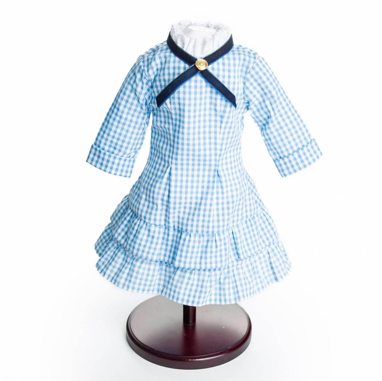 A blue and white checkered pioneer dress with a blue cross-ribbon collar and ruffled white neckline is displayed on a dark wooden stand, by The Queen's Treasures®.