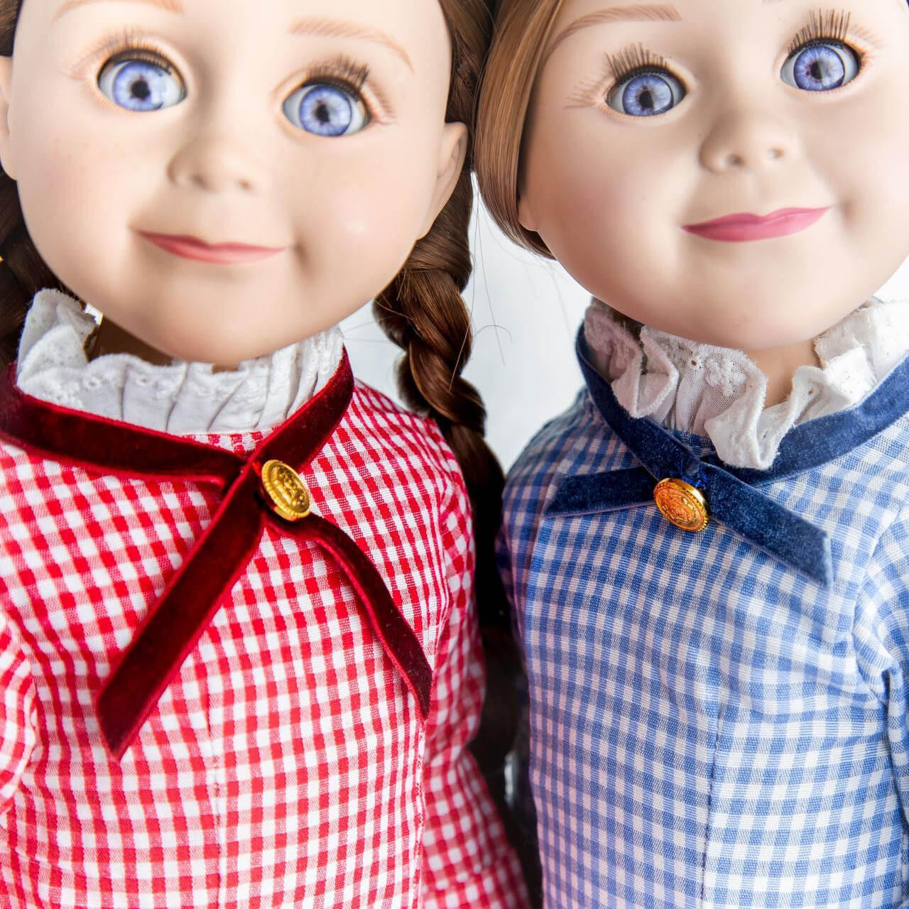 A close-up portrait of two pioneer dolls with blue eyes, one wearing a red checkered dress and the other a blue checkered dress, both featuring ruffled collars with cross-ribbon details.