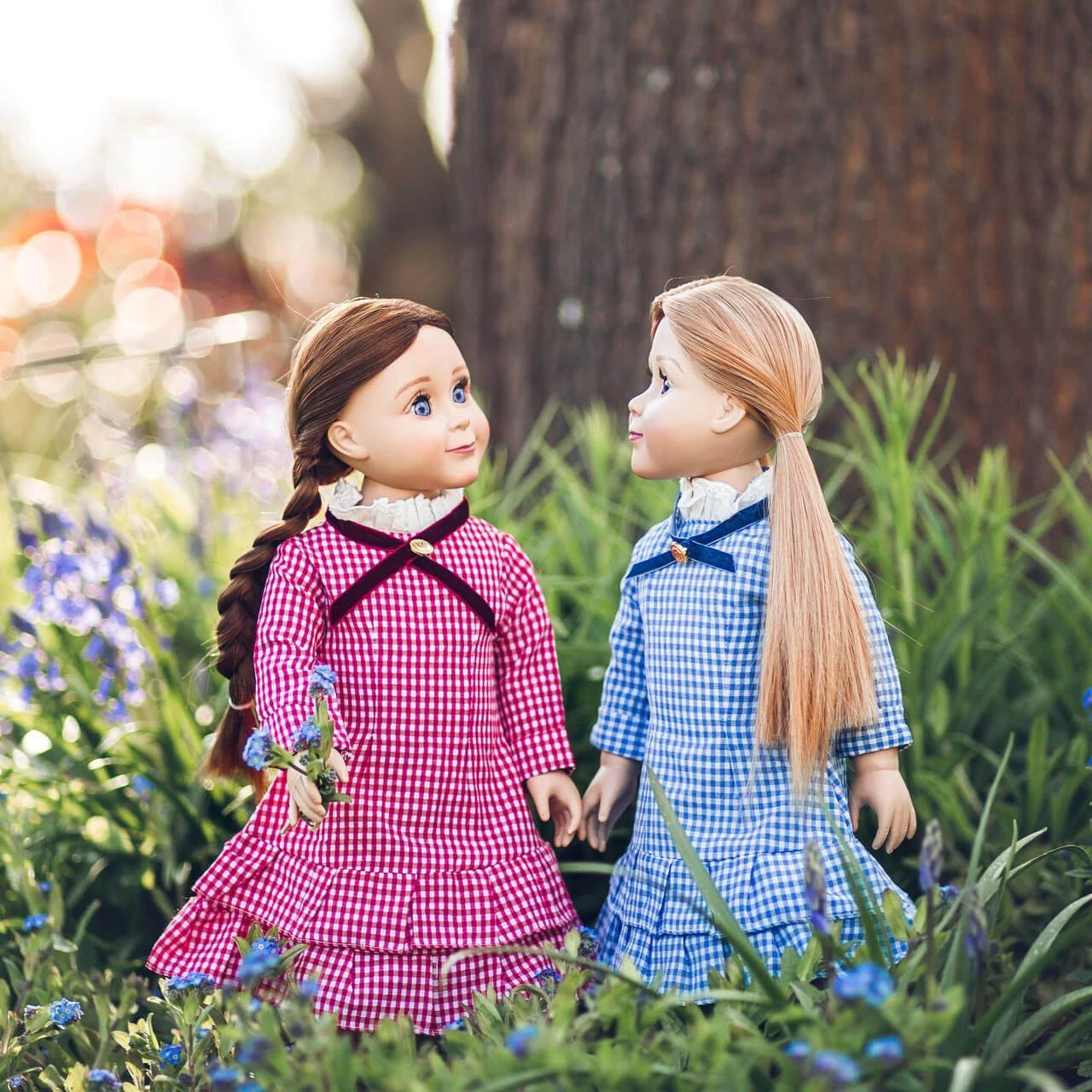 Two pioneer dolls wearing matching red and blue checkered gingham dresses stand facing each other in a grassy field next to purple wildflowers, by The Queen's Treasures®.