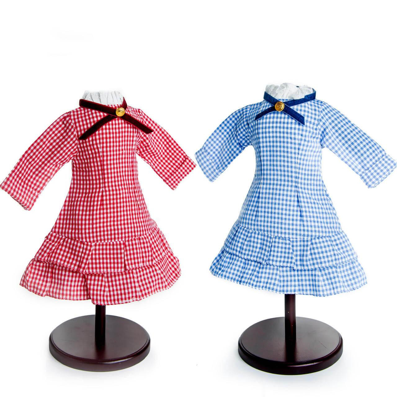 A set of two pioneer-style checkered gingham dresses, one red and one blue, are displayed on dark wooden stands against a white background, designed to fit 18" American Girl® Dolls made after 2008.