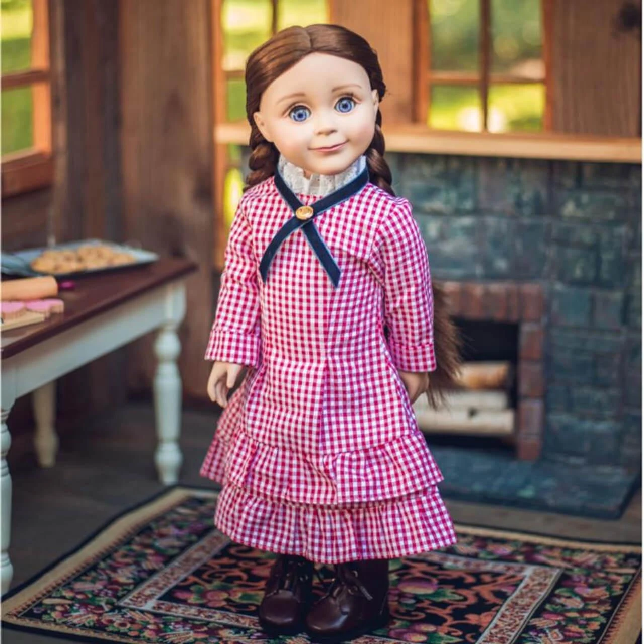 An 18-inch Little House on the Prairie® doll stands on a patterned rug inside a rustic log cabin setting. She has long brown pigtail braids and wears a red and white checkered pioneer dress featuring a high white lace collar with a dark red ribbon tie. A stone fireplace sits behind her on the right, and a white wooden kitchen table is partially visible on the left.