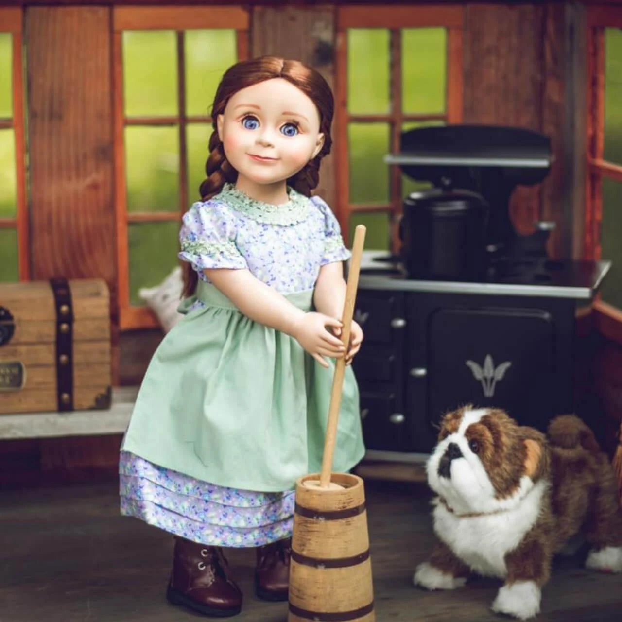 An 18-inch pioneer doll using a wooden butter churn next to a plush bulldog inside a cabin.