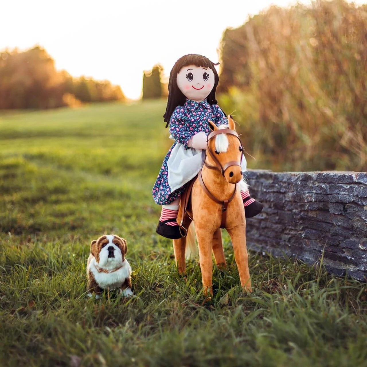 A pioneer rag doll riding a toy horse in a grassy field with a small plush bulldog sitting nearby.