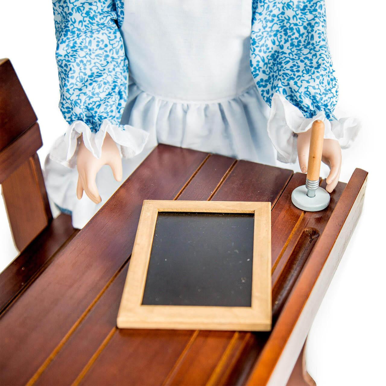 An 18-inch doll in a blue calico dress sitting at a wooden school desk with a miniature slate board with an ink well and pen.