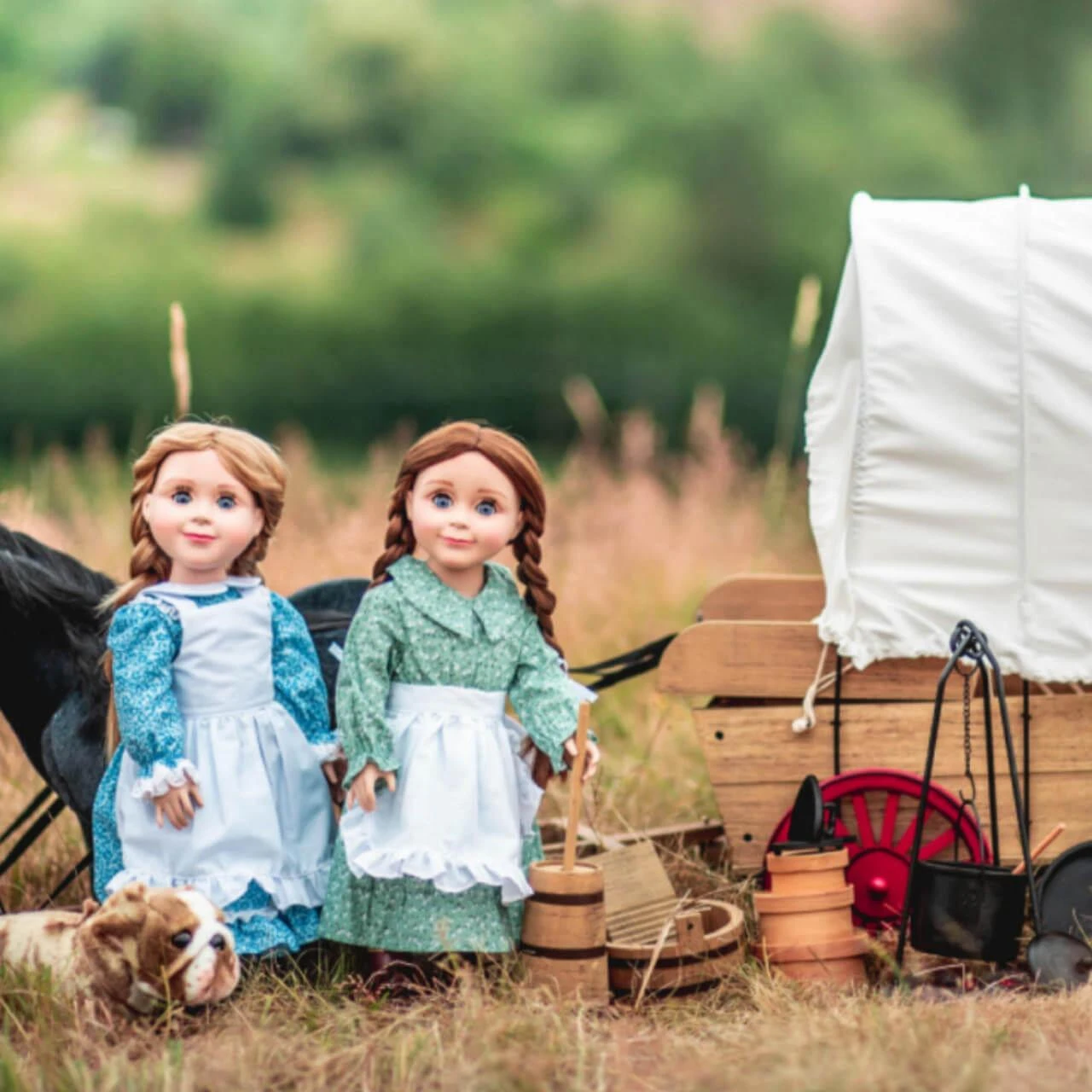 Two 18-inch pioneer dolls stand in a grassy field next to a miniature covered wagon, campfire accessories, and a small plush dog.