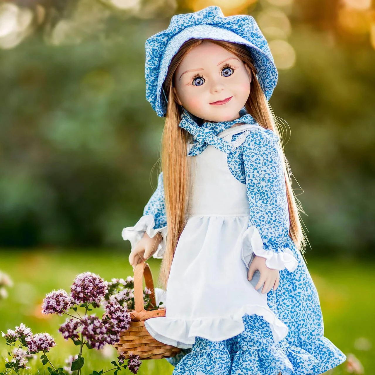 An 18-inch pioneer doll stands outdoors next to purple flowers, wearing a blue calico dress, matching bonnet, and white apron while carrying a woven basket.