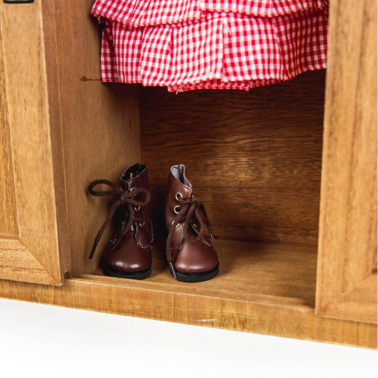 A close-up view of the bottom shelf inside a wooden doll storage trunk, showing a pair of brown lace-up boots sitting below a red gingham dress.