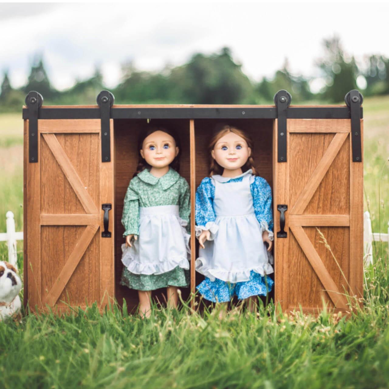 Two 18-inch pioneer dolls from Little House on the Prairie® stand inside a wooden doll storage trunk with sliding barn doors in a grassy field.