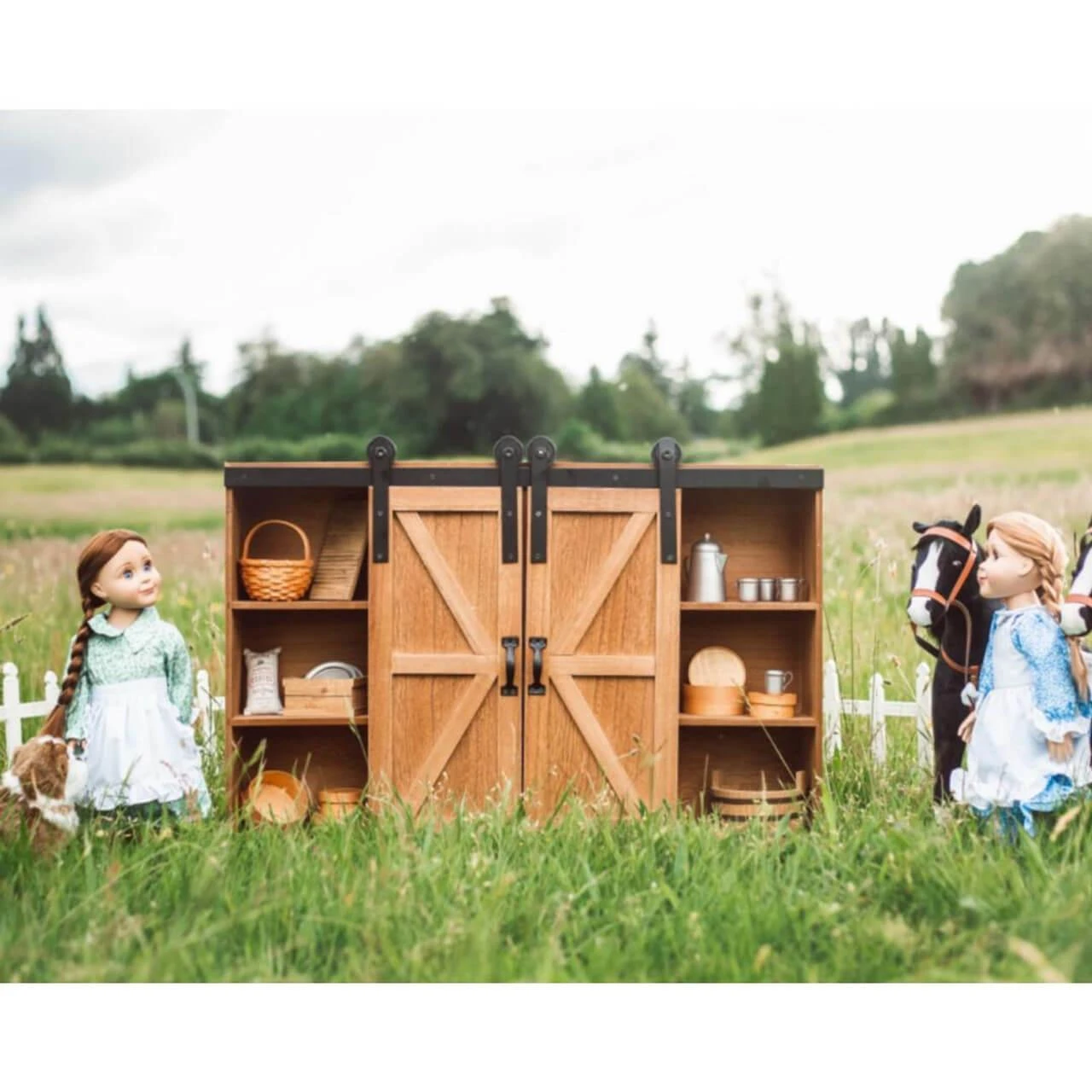 Two 18-inch pioneer dolls stand on either side of a large wooden barn-door storage trunk filled with miniature accessories in a grassy field.