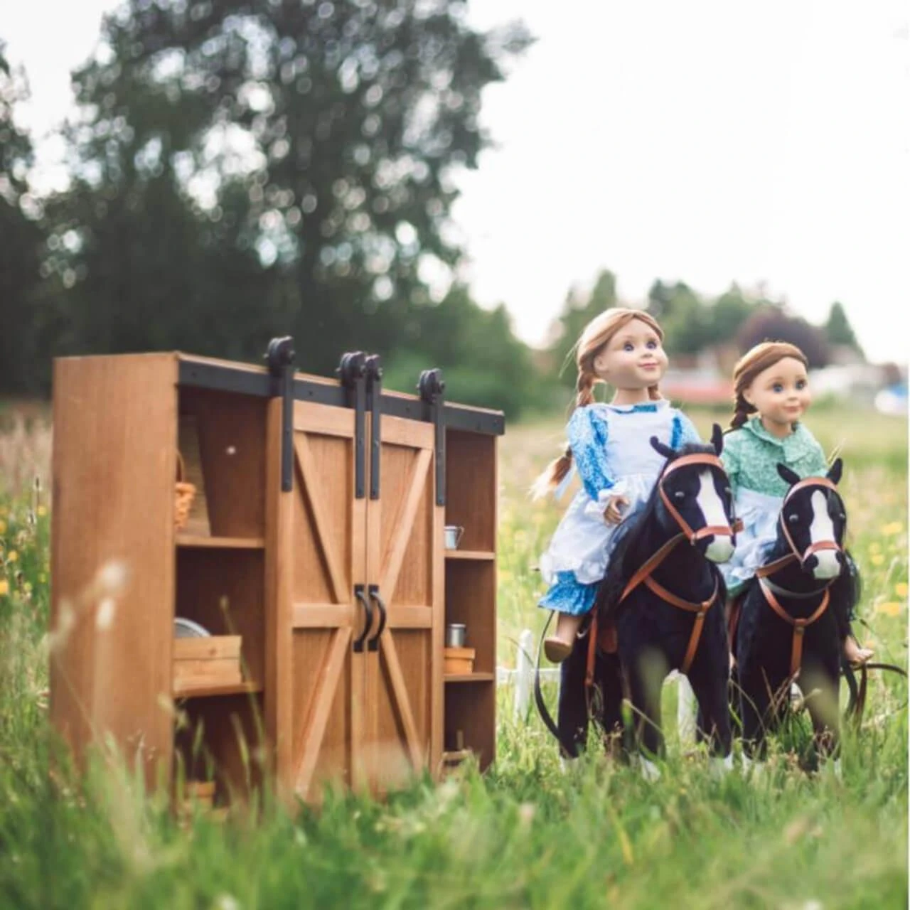 Two 18-inch pioneer dolls sit in a miniature horse-drawn carriage next to a wooden barn-door storage trunk in a grassy field.