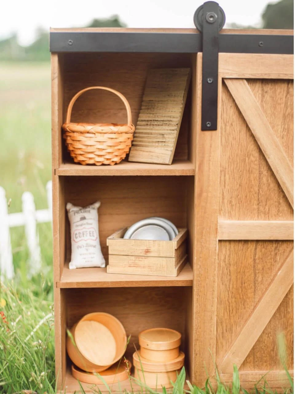A close-up view of the side shelves of a wooden doll storage trunk, filled with miniature pioneer accessories including a basket, a washboard, plates, and small wooden containers.