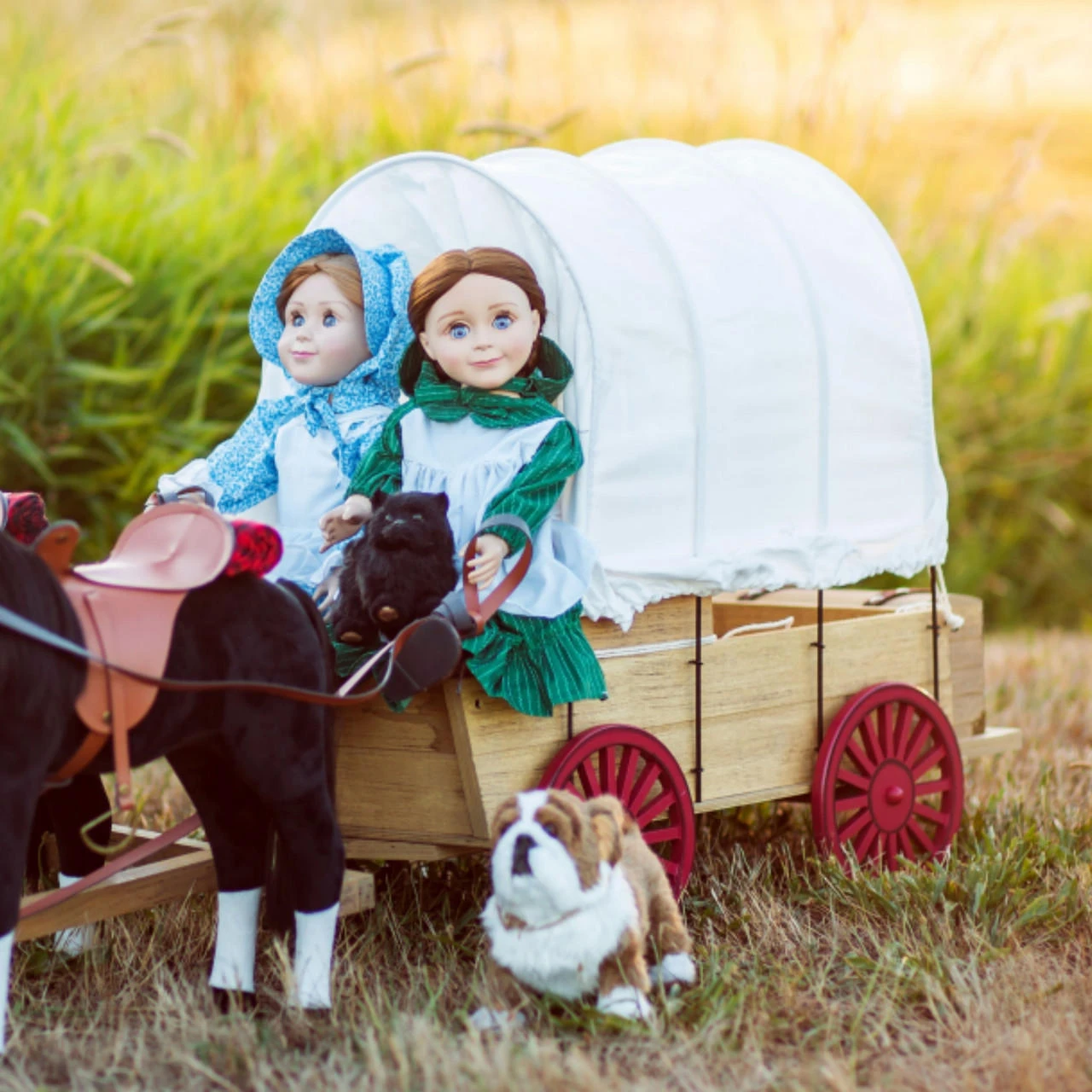 Two 18-inch pioneer dolls sit inside a wooden covered wagon with a white fabric canvas and red wheels from the Little House on the Prairie® collection, hitched to a black toy horse while a plush bulldog sits nearby in a grassy field.