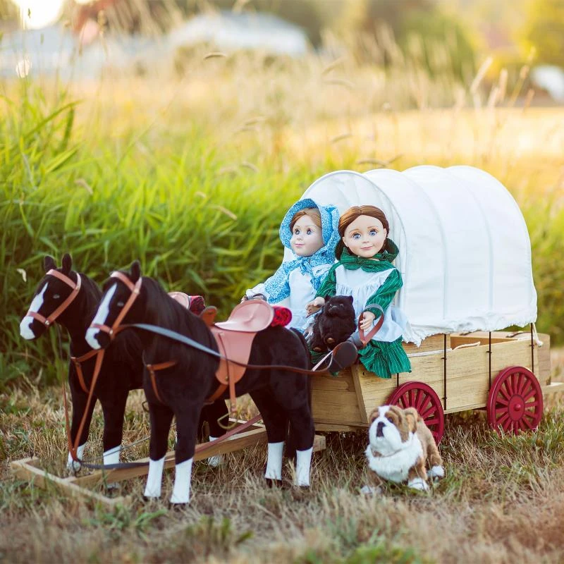 Two 18-inch dolls by The Queens Treasures® sit in a wooden covered wagon pulled by a team of two black horses, accompanied by a small plush bulldog in a sunlit grassy field.