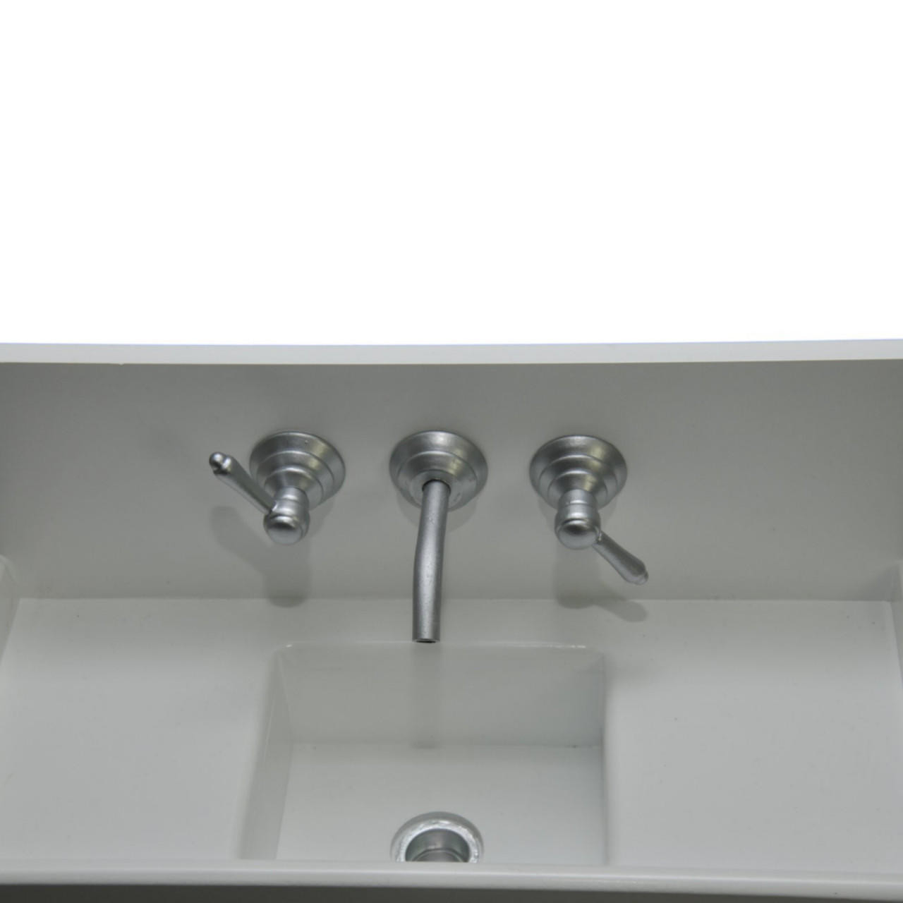 A top-down view shows a miniature white vintage-style farmhouse kitchen sink against a solid white background. The empty doll-sized sink features a rectangular deep central basin with a small silver drain at the bottom, a flat top deck, and a silver three-piece faux wall faucet with a central downward-curved spout and two matching lever handles mounted on the backsplash.