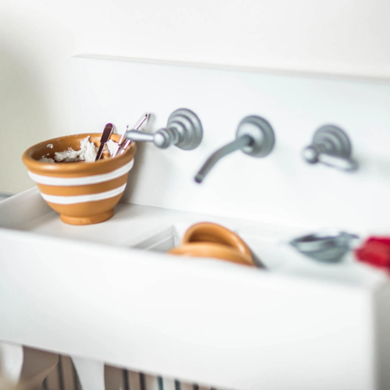 A close-up view shows a miniature white farmhouse-style toy kitchen sink filled with tiny dishes. Mounted on the high backsplash is a grey three-piece faux wall faucet with a central spout and two side lever handles. Inside the shallow rectangular basin rests a yellow and white striped mixing bowl holding miniature utensils, a yellow dish, and a grey handheld tool with a red handle.