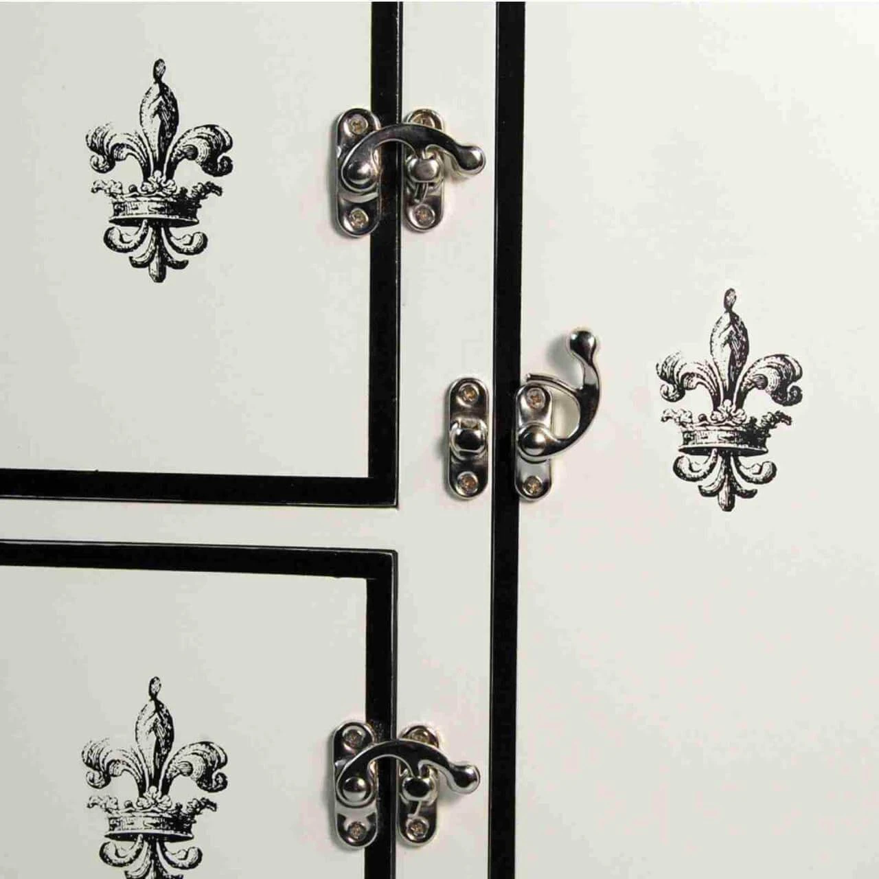 A close-up view shows the decorative details on a miniature white vintage icebox cabinet. Three polished silver-colored metal latches secure the cabinet doors along thin black painted trim borders. A black-and-silver ornate fleur-de-lis emblem with a crown motif is centered on each visible door panel.