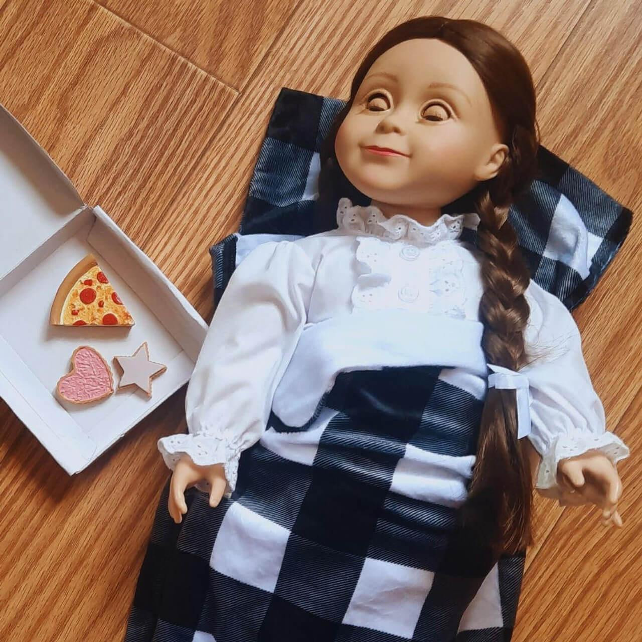 An angled overhead photograph shows an 18-inch doll tucked into a miniature plaid sleeping bag next to tiny toy treats on a hardwood floor.