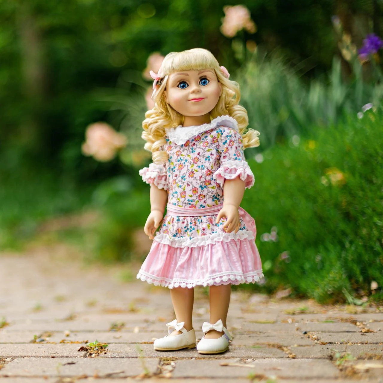 An 18-inch pioneer doll with curly blonde hair stands on a brick path outdoors, wearing a pink floral calico dress with lace ruffles.