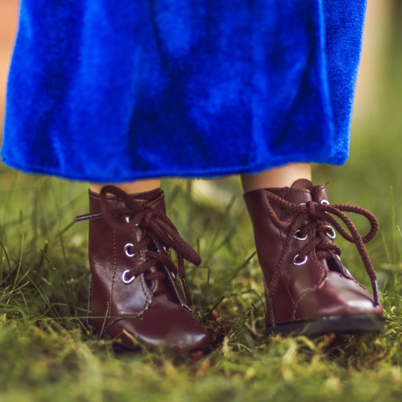 A close-up shot focuses on the lower legs of an 18-inch doll wearing a vibrant, bright blue velvet dress. The doll stands outdoors on green grass, showcasing a pair of miniature dark brown, glossy lace-up boots. The boots feature thick matching brown laces tied in bows and are detailed with small silver eyelets.