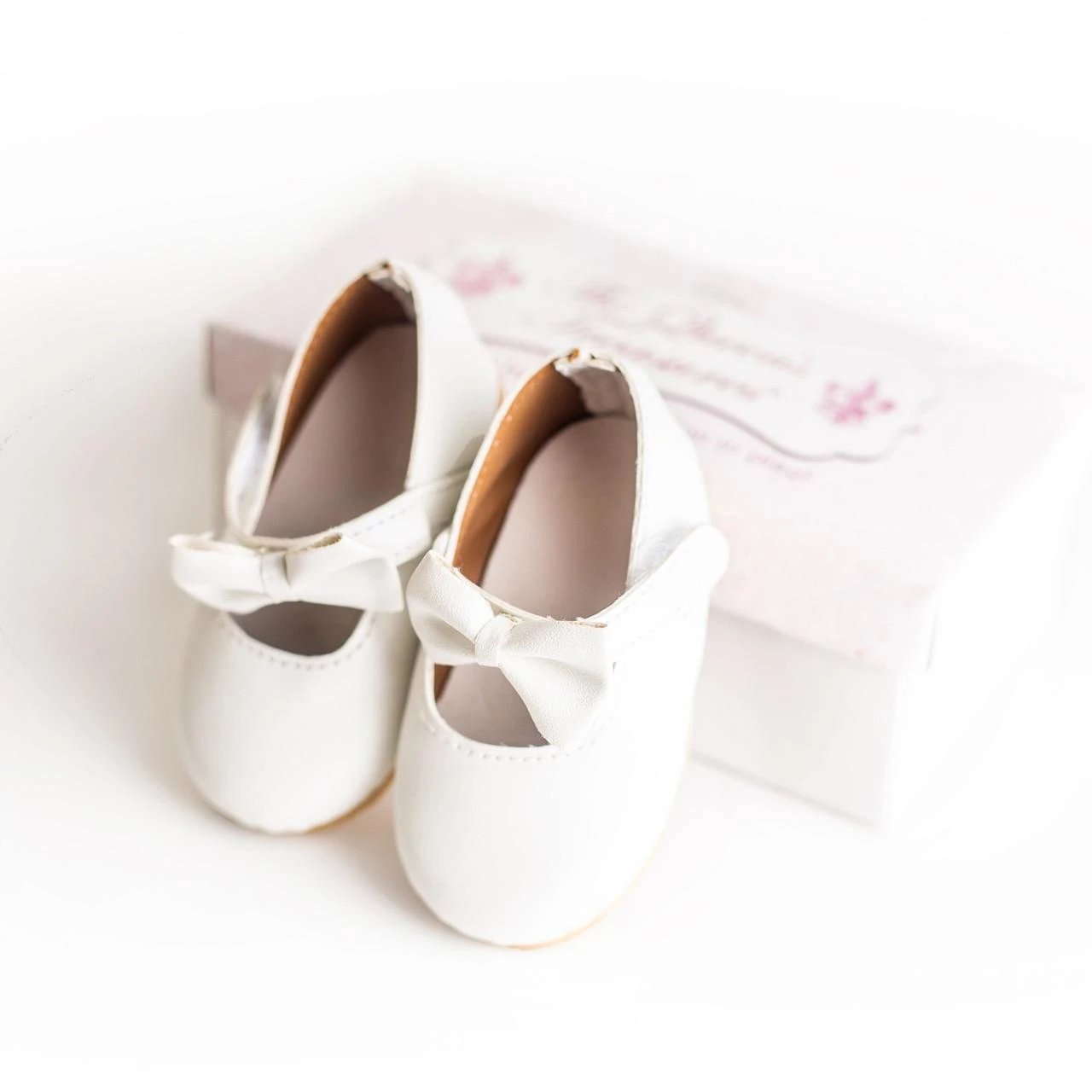 A pair of miniature white Mary Jane toy shoes sits upright against a plain white background. The shoes feature a smooth white finish, light tan insoles, and a matching white bow accent detailed on each front strap. A white rectangular product box with subtle pink and grey graphics is positioned slightly behind and to the right of the shoes.