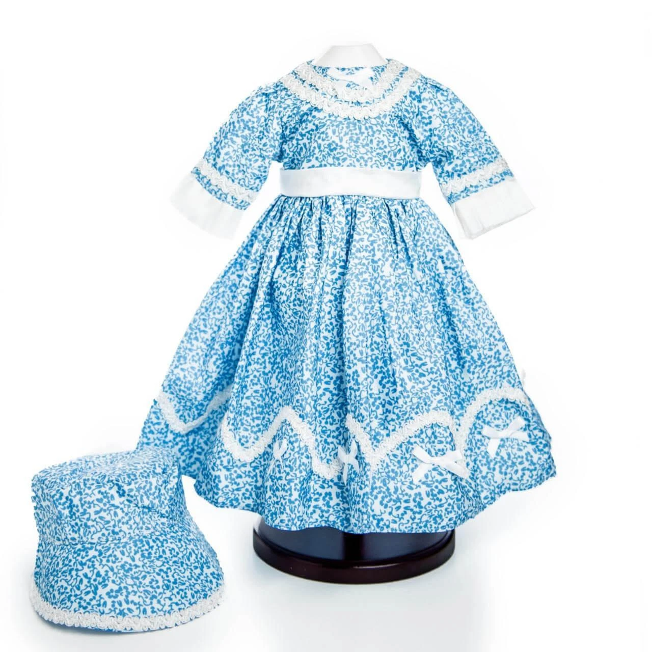 A historical-style blue and white floral patterned doll dress is displayed on a black stand next to a matching bonnet against a plain white background. The long-sleeved dress features white lace detailing around the high collar, white fabric cuffs, a white waistband, and a wavy white lace trim near the hem of the full skirt. The matching bonnet sits to the lower left of the dress stand.