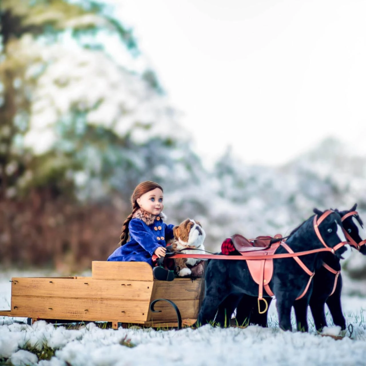 An 18-inch doll with brown hair braided into pig-tails sits inside a wooden toy sleigh pulled by a pair of black toy horses in a snowy, outdoor setting. The doll is wearing a wintery royal blue coat with brown faux-fur trim. A small plush bulldog toy sits on the bench seat right next to her. The two toy horses are outfitted with detailed brown saddles and red harnesses, all positioned on snow-dusted grass with blurry pine trees in the background.