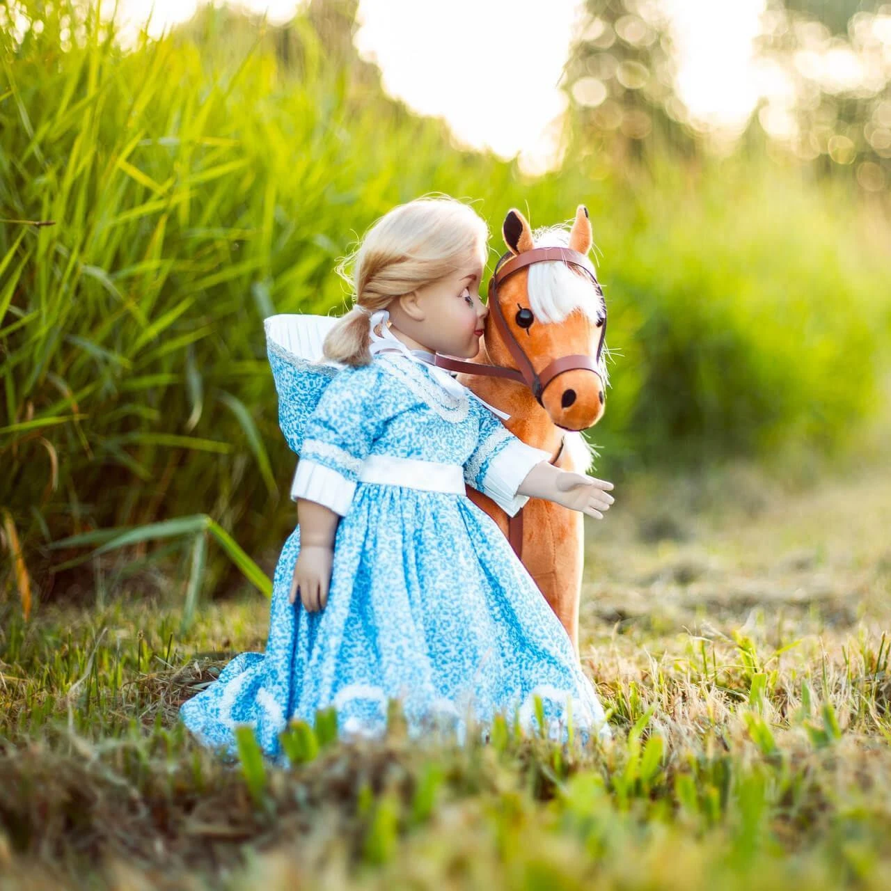 An 18-inch doll with blonde hair stands outdoors in a grassy field next to a toy pony. The doll wears a historical-style blue and white floral dress with white trim at the collar, waist, and cuffs, paired with a matching blue floral bonnet. She stands close to a tan toy horse that has a white patch on its face and a brown bridle, set against a background of tall green grass illuminated by soft sunlight.