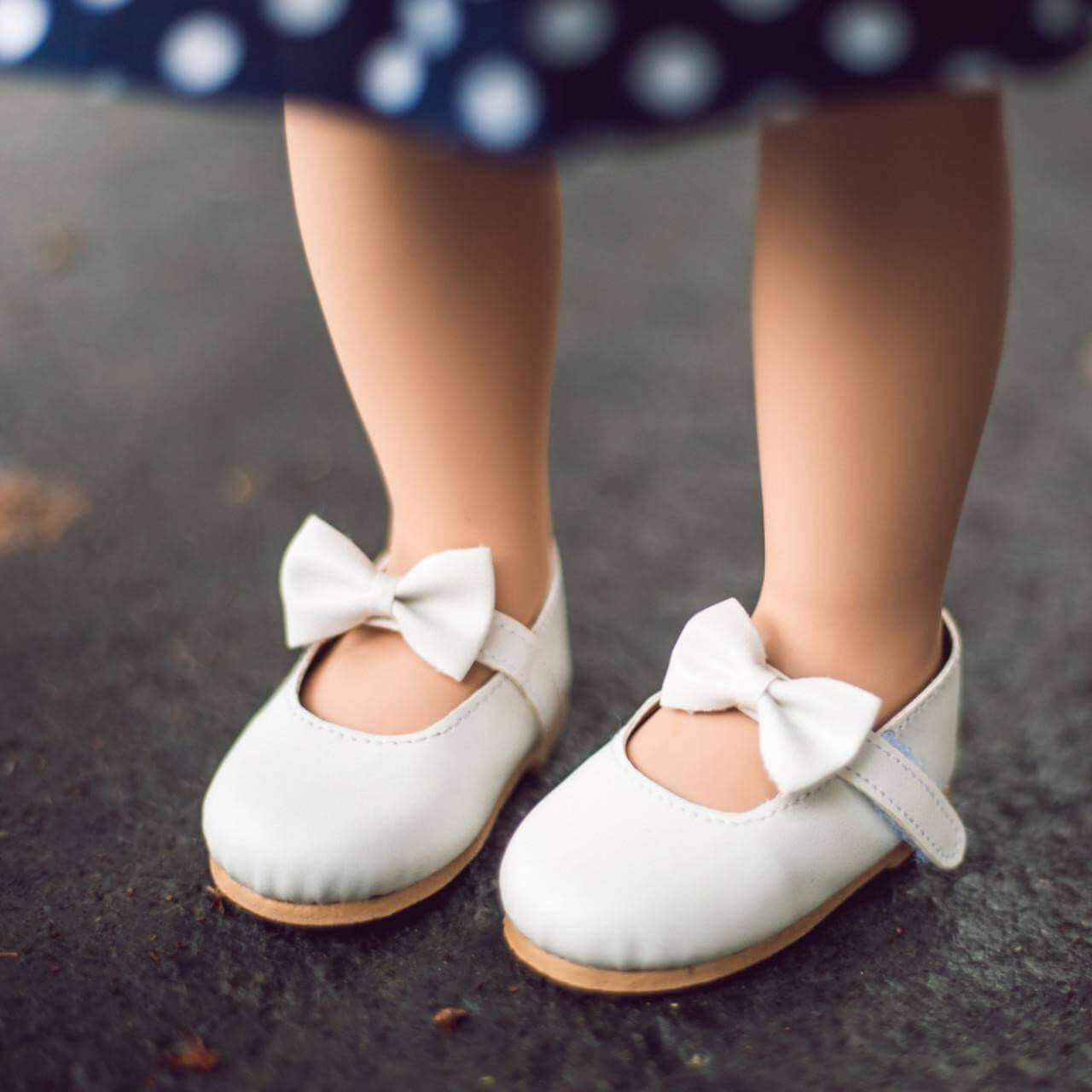A close-up photograph shows an 18-inch doll wearing white dress shoes while standing on a textured grey surface.