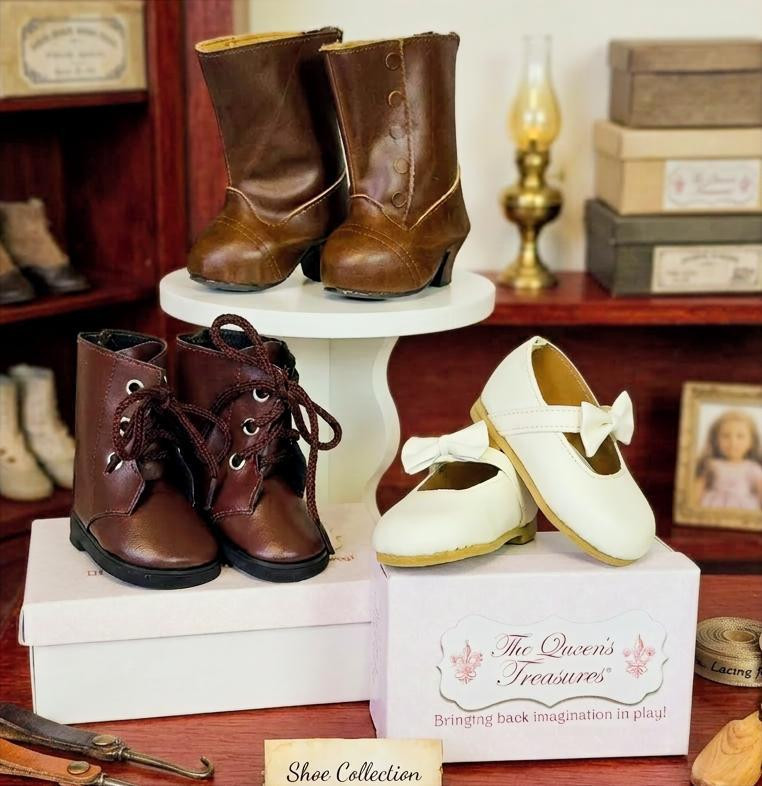 Three distinct styles of miniature footwear designed to fit 18-inch American Girl® dolls.  Tall Riding Boots: A pair of tall, glossy brown pull-on boots displayed prominently on a small white pedestal stand. Lace-Up Ankle Boots: A pair of dark brown ankle boots with realistic eyelets and tied laces resting on a white rectangular box. Mary Jane Flats: A pair of classic cream-colored Mary Jane shoes with small decorative bows on the straps leaning against a white branded shoe box. This is inside a doll shop setting.