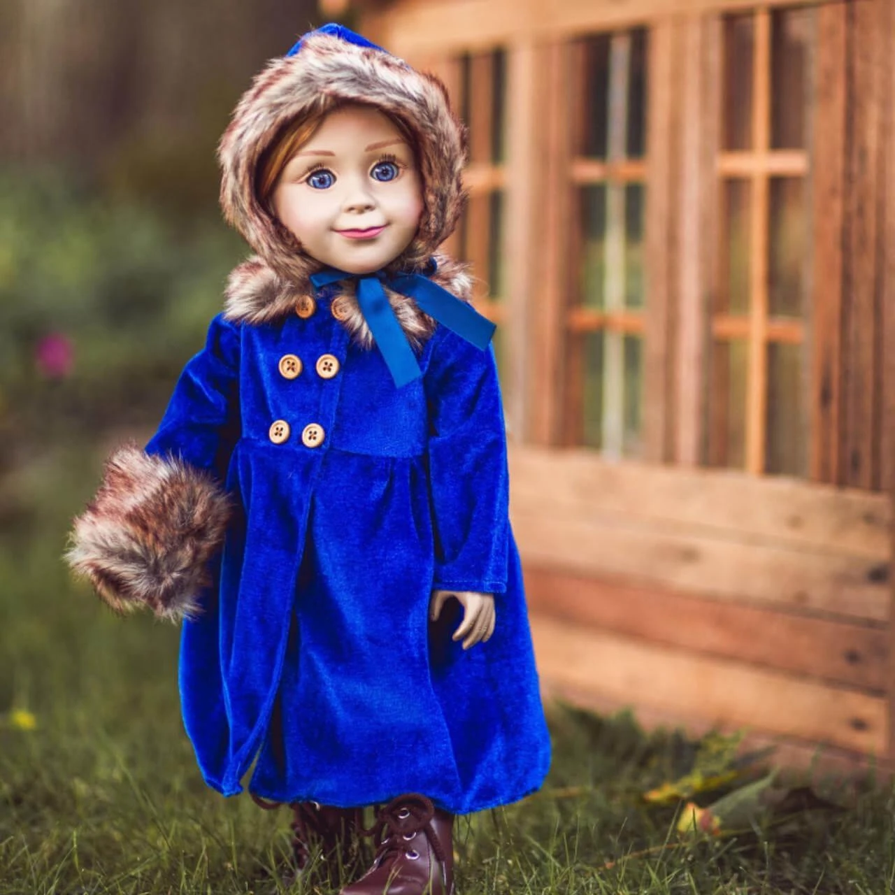 An 18-inch doll stands outdoors on the grass in a royal blue velvet winter coat with four gold buttons, a matching brown faux fur hat tied with a blue ribbon, and a faux fur hand muff, set against a blurred wooden house background.