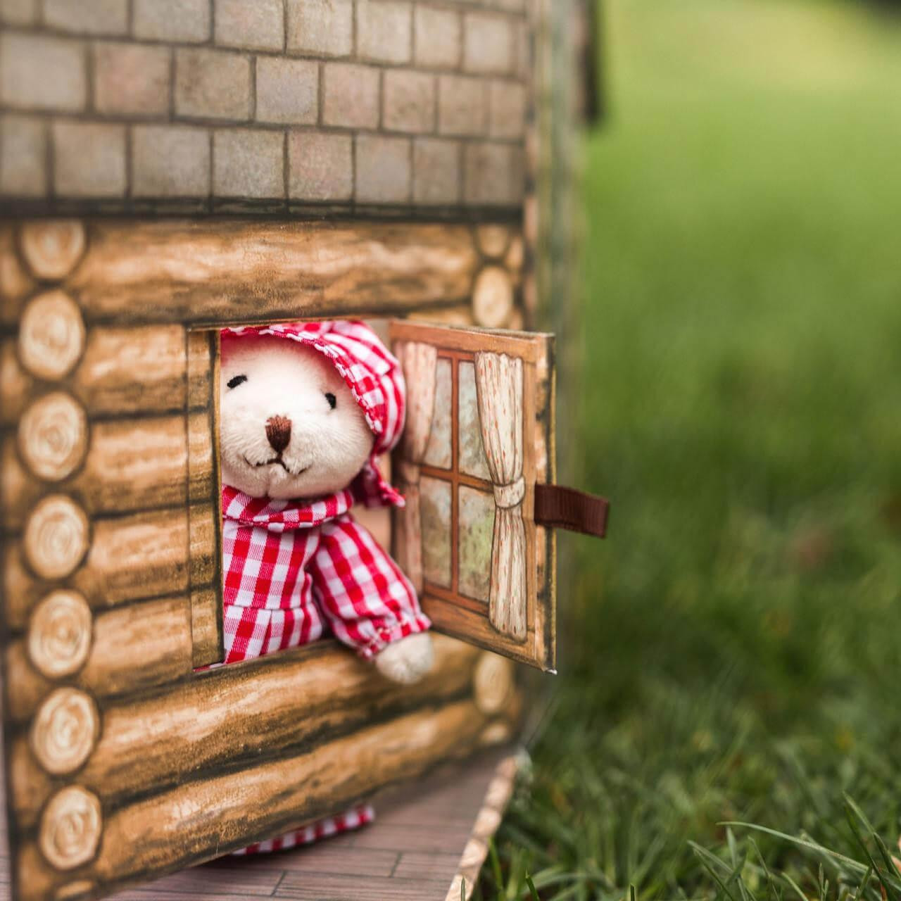 A small plush teddy bear in red and white checkered dress looks out from the open window of a toy log cabin playset outdoors against a blurred green grass background.