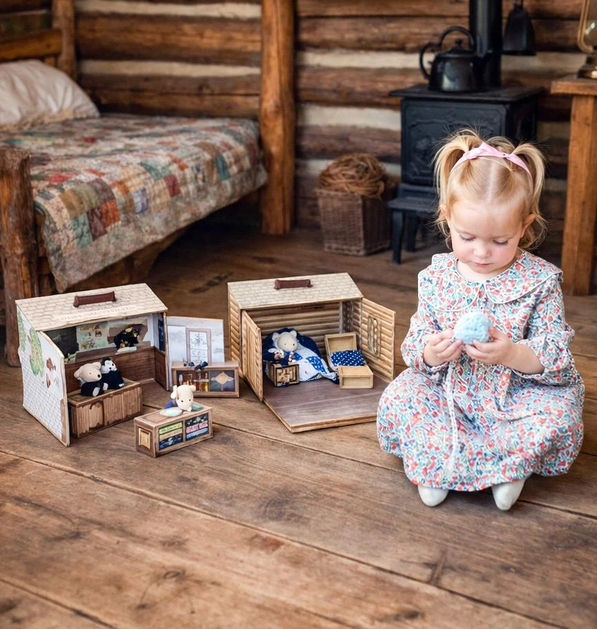 Two open, miniature pop-up suitcase playsets depicting scenes from Little House on the Prairie® are displayed against a white background, filled with small plush teddy bears dressed in period clothing amidst detailed general store and log cabin backdrops. These Little House on the Prairie toys are inside a cabin with a little blonde child playing with it