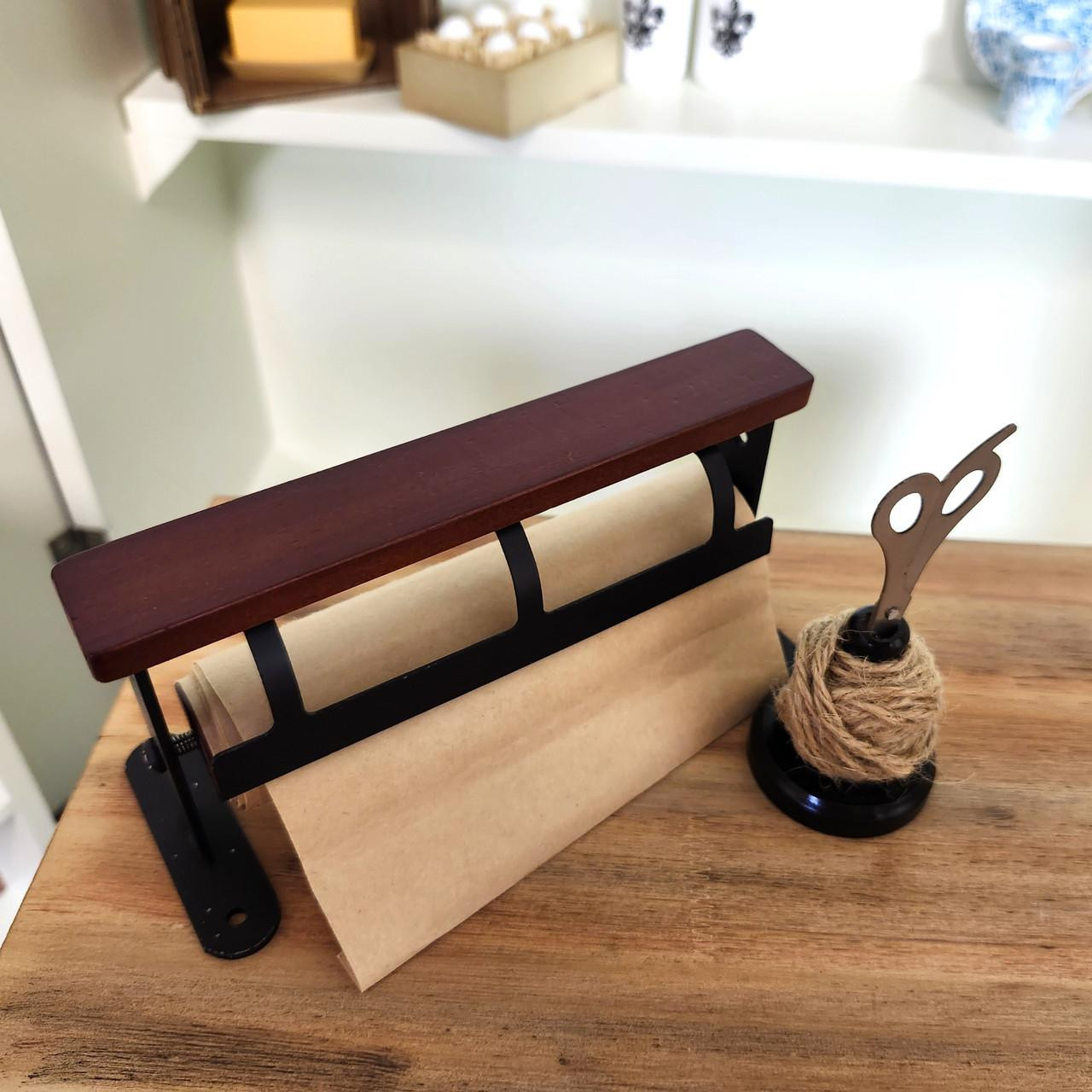 A miniature wooden wrapping paper dispenser with tan paper alongside a matching ball of twine and tiny scissors from the Little House on the Prairie® collection, displayed on a wooden surface.