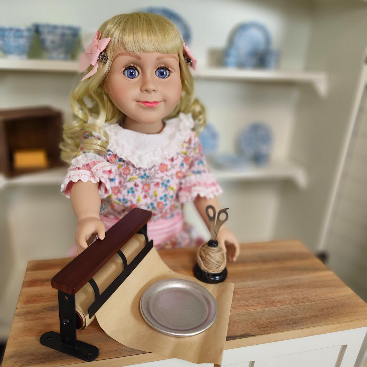 An 18-inch doll stands behind a wooden counter by The Queens Treasures®, using a miniature vintage wrapping paper dispenser with brown paper, a tiny metal plate, and a small ball of twine with scissors.