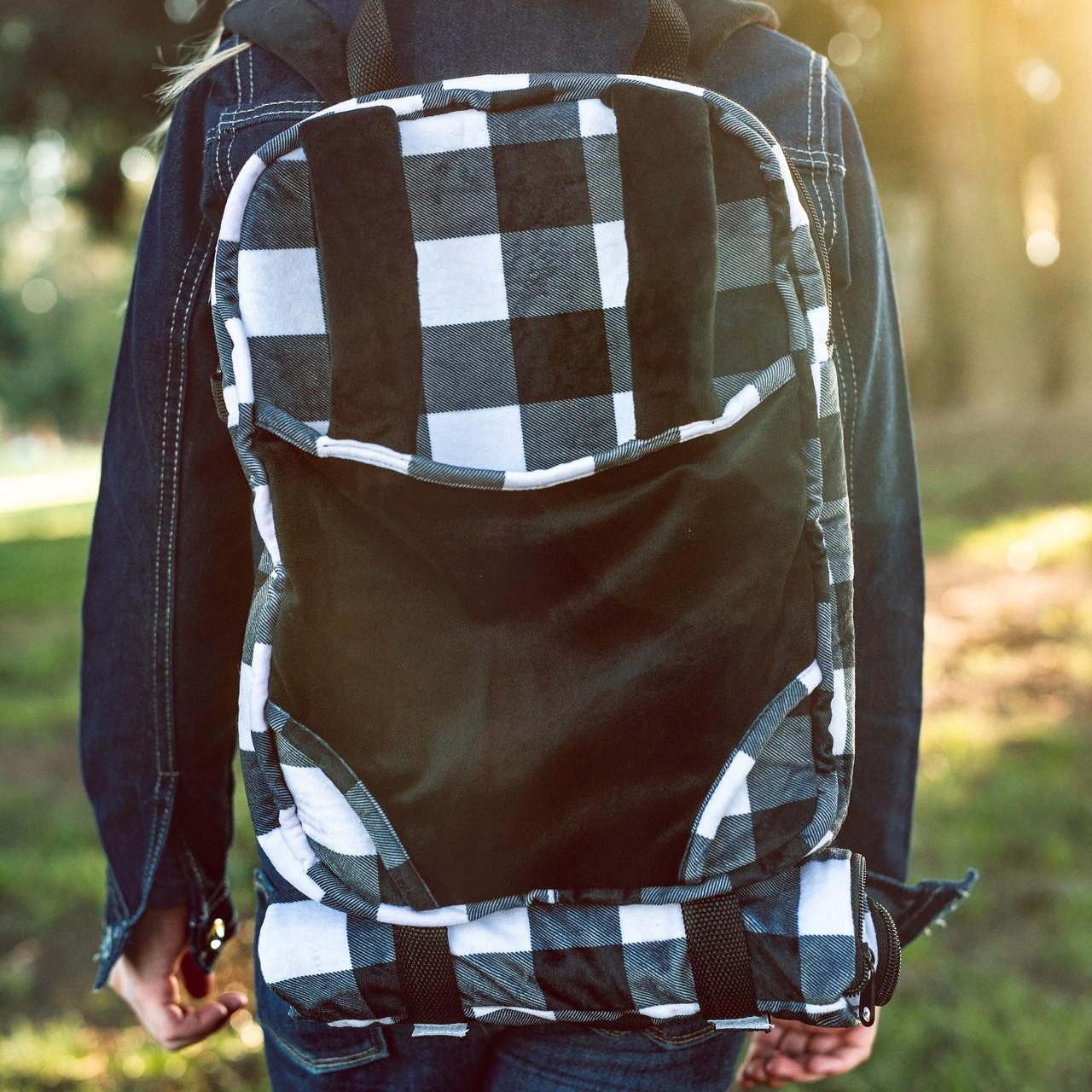 A person viewed from behind wears a plush doll-carrier backpack featuring a black and white buffalo plaid pattern. The backpack includes a solid black front panel, matching plaid shoulder straps, and a rolled-up buffalo plaid sleeping bag secured horizontally across the bottom while outdoors.