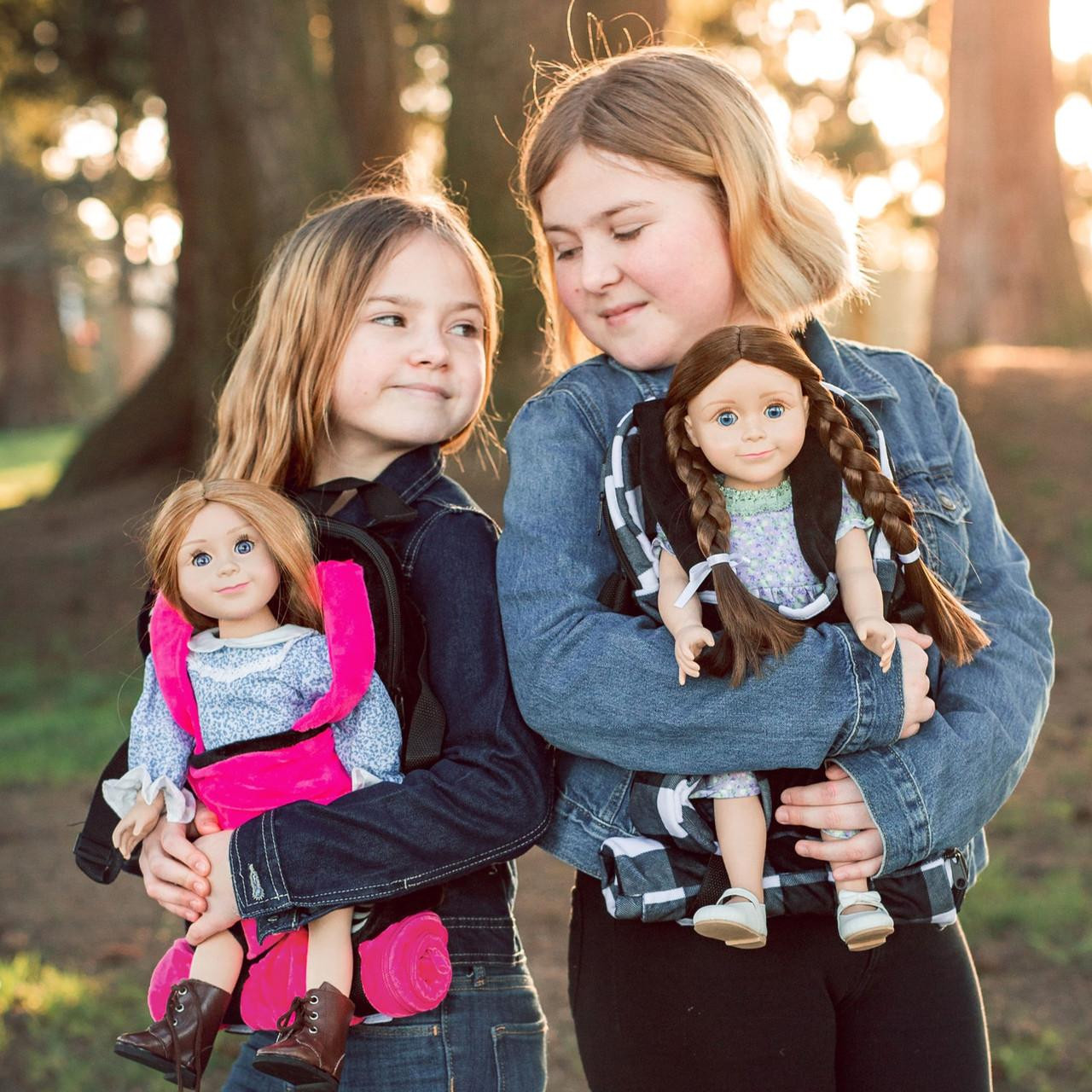 Two young girls stand close together outdoors in a wooded setting, each holding a doll tucked into a specialized carrier backpack. The younger girl on the left wears a dark jacket and turns her head toward the older girl, holding a doll with strawberry-blonde hair in a vibrant pink carrier. The older girl on the right wears a denim jacket and smiles gently down at her doll, which features long brown braids and sits securely in a black-and-white buffalo plaid carrier.