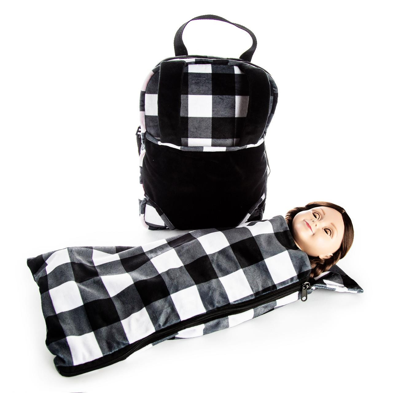 A plush doll-carrier backpack sits upright next to a doll tucked inside a matching sleeping bag against a plain white background. The backpack features a solid black lower panel and straps over a black and white buffalo plaid upper section. The doll, with dark hair, rests on its back with only its head visible from the zipped-up buffalo plaid sleeping bag. Carrier is 10"W x 3"D x 15"H and the sleeping bag measures 10" x 19". Perfect fit for your 18 inch dolls.