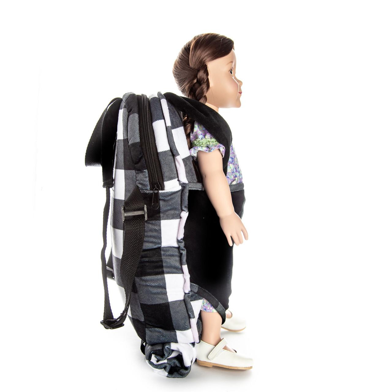A side-profile view of a doll secured inside a plush doll-carrier backpack featuring a black and white buffalo plaid pattern. The doll faces right, dressed in a patterned dress and white shoes, with a matching black front carrier and a rolled-up plaid sleeping bag visible underneath against a plain white background.