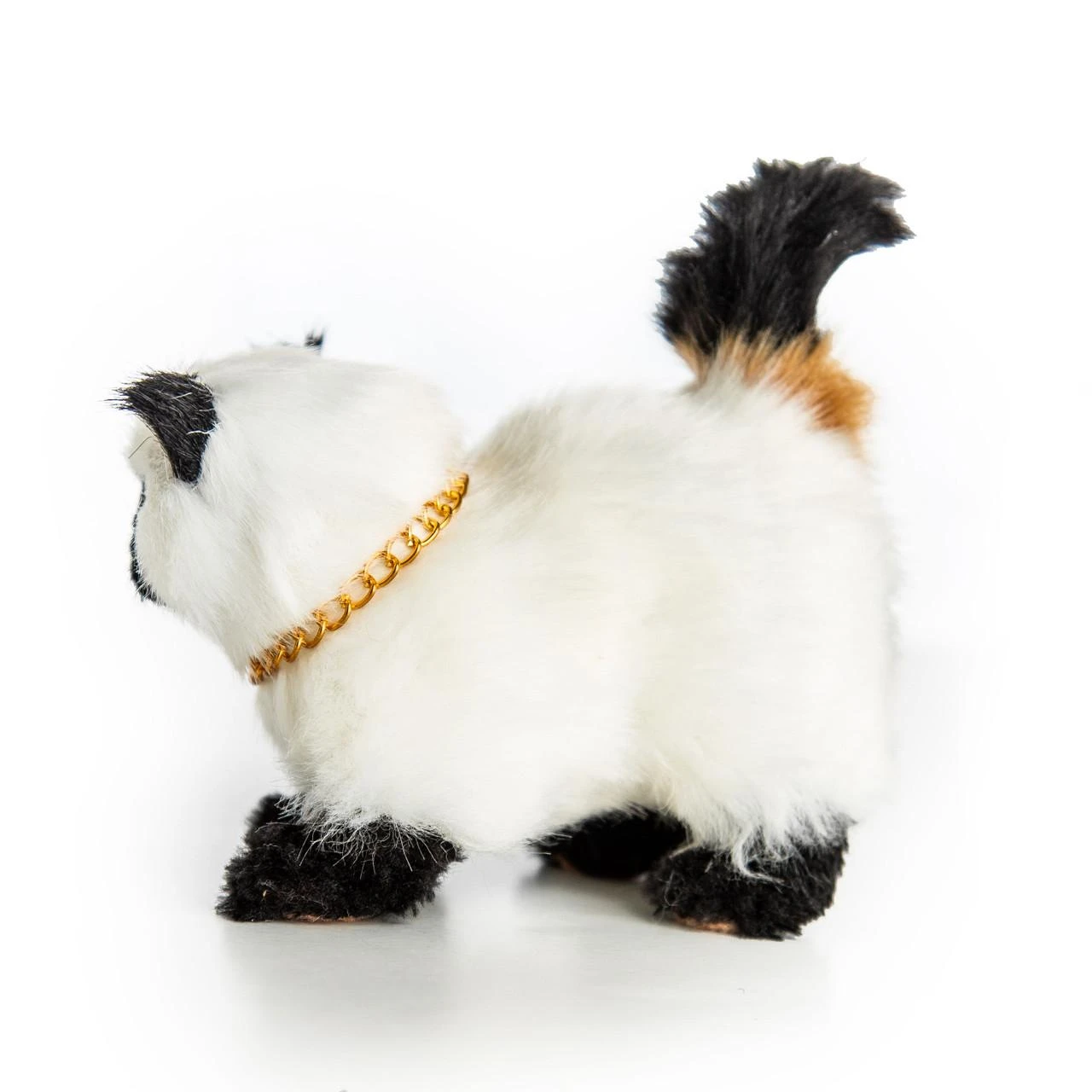 A realistic, fluffy Siamese-style toy cat is viewed from behind as it walks toward the left against a plain white background. The toy cat features a cream-colored plush body with black ears, black paws, a gold link chain collar, and a raised black tail with a patch of tan fur at its base.