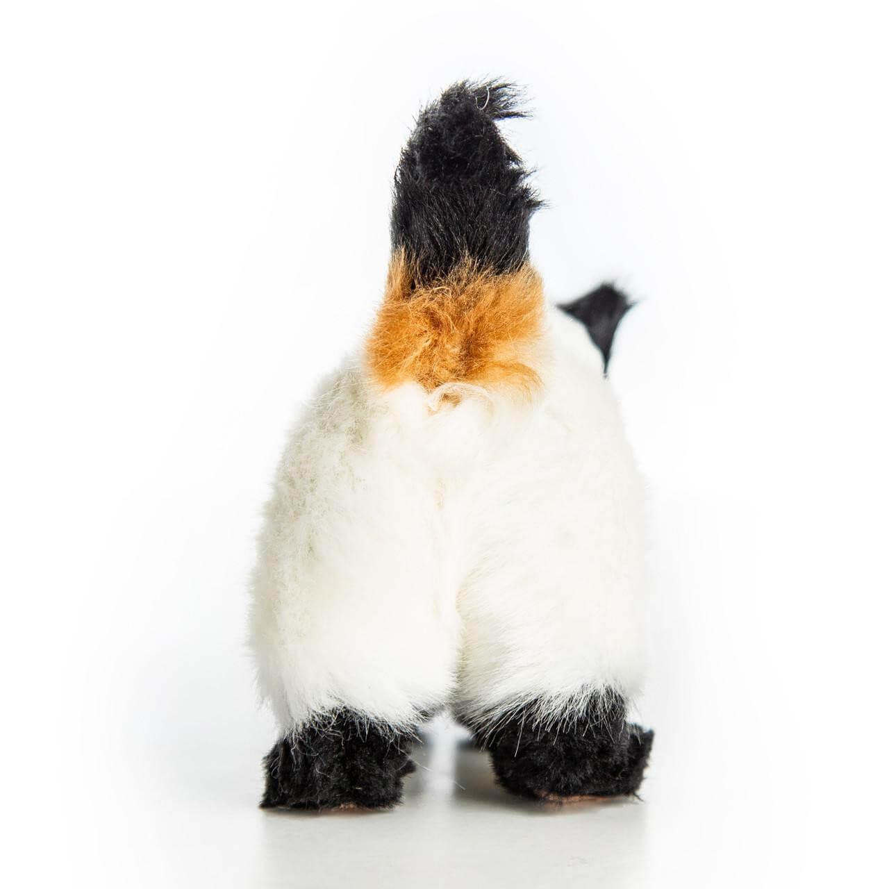 A rear view of a realistic, fluffy Siamese-style toy cat standing on a plain white background. The toy cat has a cream-colored body, black paws, and a prominent upright tail that features a bright orange-tan patch at its base and a black tip.