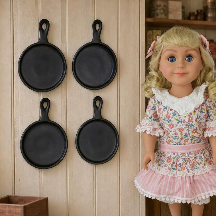 An 18" Doll stands by a wooden wall displaying four miniature black cast iron-style frying pans from the Little House on the Prairie® collection by The Queens Treasures®.
